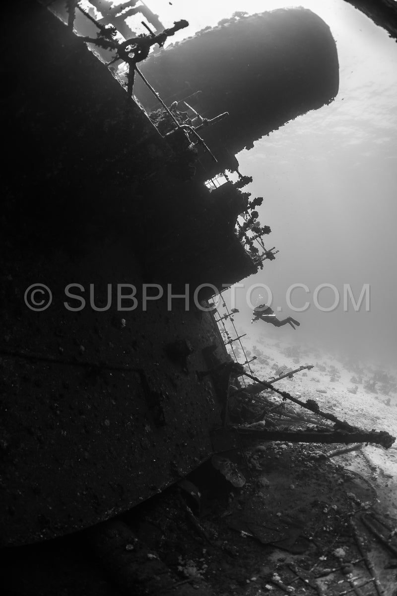 A diver- using a rebreather- swims along the side of a shipwreck in the Red Sea. The diver is silhouetted against the sunlight- making their figure dark against the lighter background.