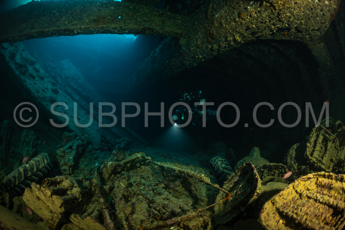 A diver wearing a rebreather explores the interior of a shipwreck in the Red Sea. The diver's light illuminates the dark interior- revealing the rusted and corroded metal of the ship.