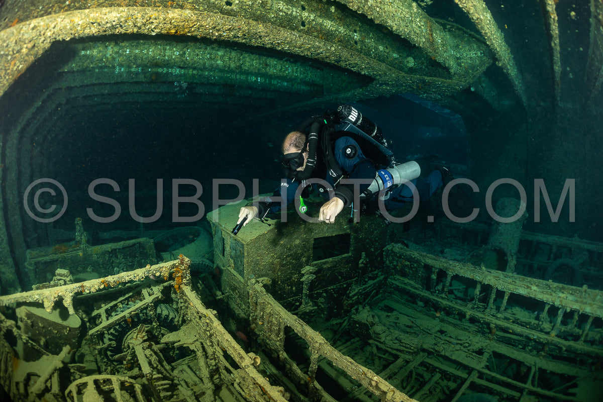 A diver wearing a rebreather swims through the interior of a shipwreck in the Red Sea. The diver is illuminated by a flashlight.