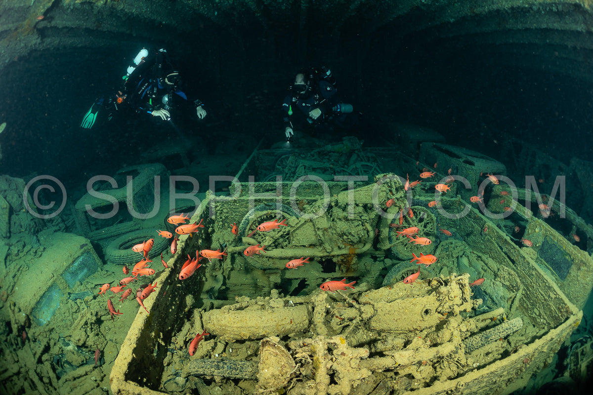 A diver wearing a rebreather explores a shipwreck in the Red Sea. The wreck is covered in marine life- including a school of red fish. The diver is silhouetted against the sunlight coming from above.