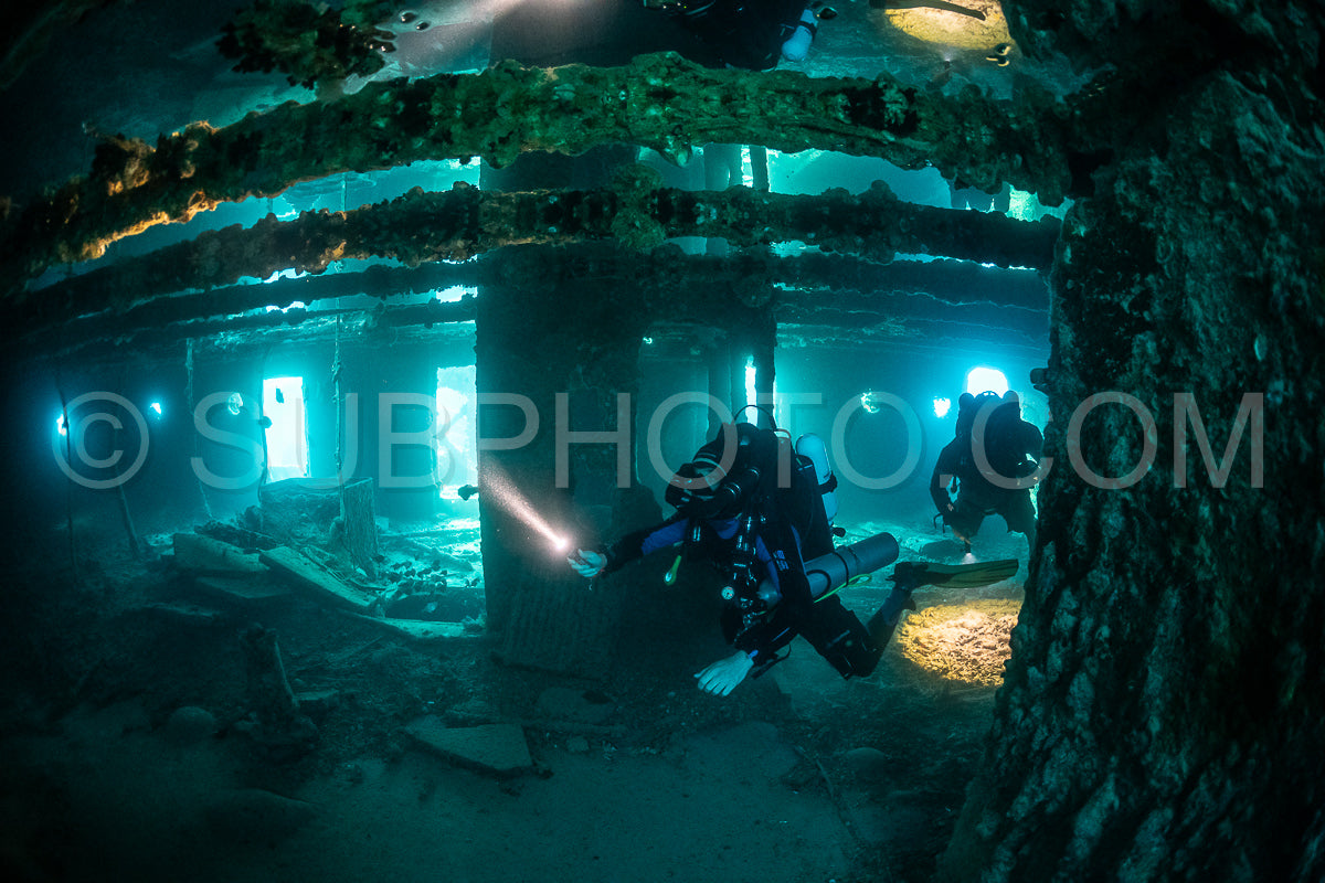 A scuba diver- wearing a rebreather and carrying a dive light- swims through a large- submerged shipwreck in the Red Sea.