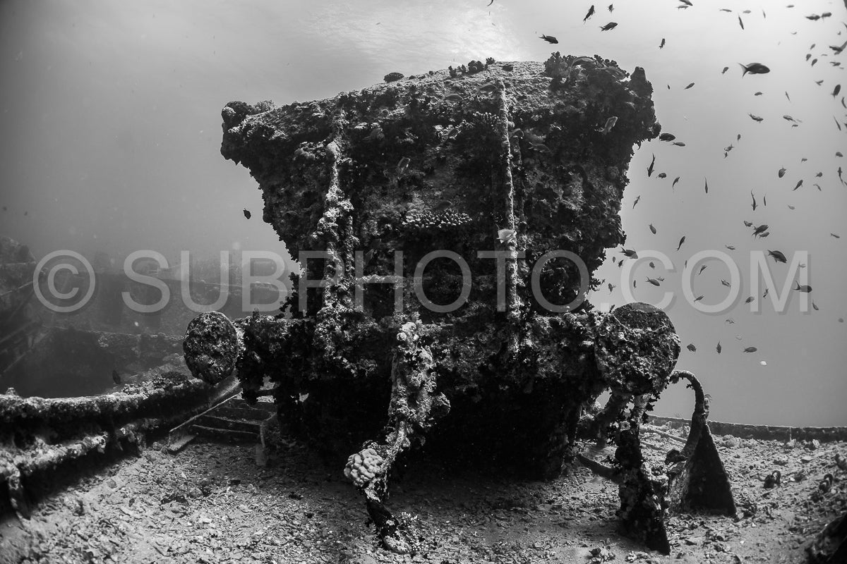 A diver wearing a rebreather is silhouetted against the metal structure of a sunken ship. The water is murky- but the diver's form is visible against the wreckage.