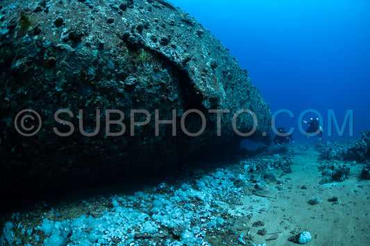 A scuba diver wearing a rebreather swims through an opening in a wreck in the Red Sea. Thewreck is covered in marine life and the ocean floor is sandy.