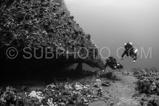 Two divers wearing rebreathers are underwater- swimming past a wreck.