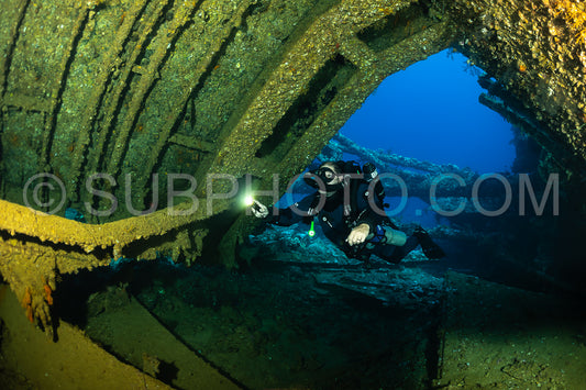 A diver with a rebreather swims through a large opening in a shipwreck- illuminating the dark interior with a flashlight.