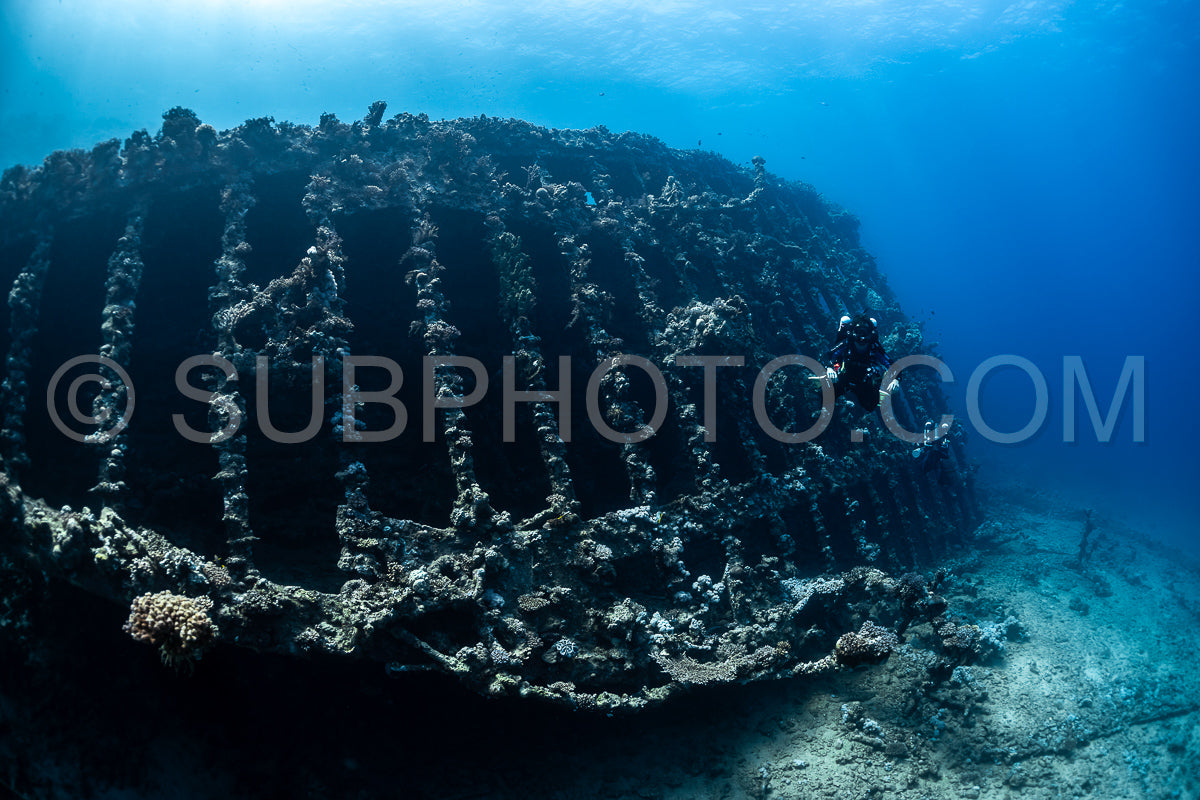 A diver wearing a closed-circuit rebreather swims along the side of a shipwreck covered in coral. The shipwreck is large and has a series of vertical beams.