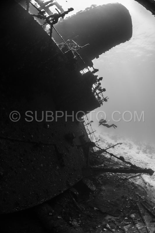 A diver- using a rebreather- swims along the side of a shipwreck in the Red Sea. The diver is silhouetted against the sunlight- making their figure dark against the lighter background.
