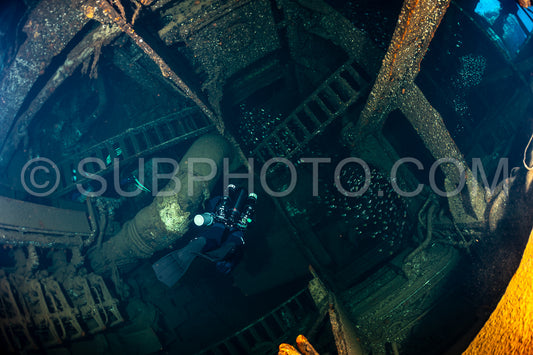A diver wearing a rebreather explores the interior of a shipwreck in the Red Sea. The diver is illuminated by a light and is surrounded by the dark- rusty metal of the sunken vessel.