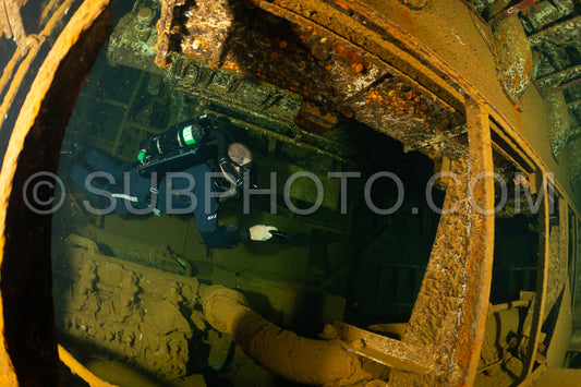 A diver wearing a rebreather swims through a narrow opening inside a sunken shipwreck in the Red Sea. The metal hull of the ship is covered in rust and marine life.