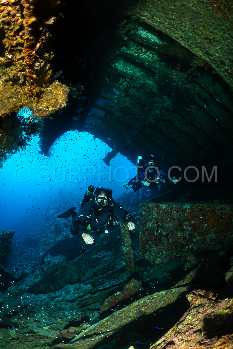 Two divers are exploring the interior of a shipwreck in the Red Sea. The diver in the foreground is using a rebreather to explore the wreck.