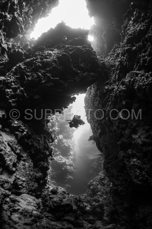 A diver is silhouetted against a beam of sunlight as they pass through a narrow gap in a coral reef. The diver is using a rebreather and is surrounded by dark- rough coral formations.