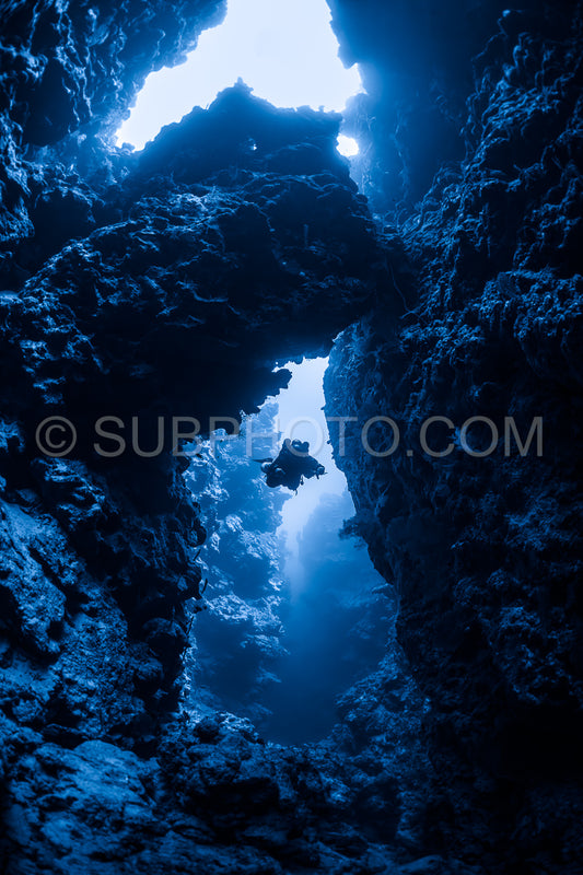 A diver is silhouetted against a beam of sunlight as they pass through a narrow gap in a coral reef. The diver is using a rebreather and is surrounded by dark- rough coral formations.