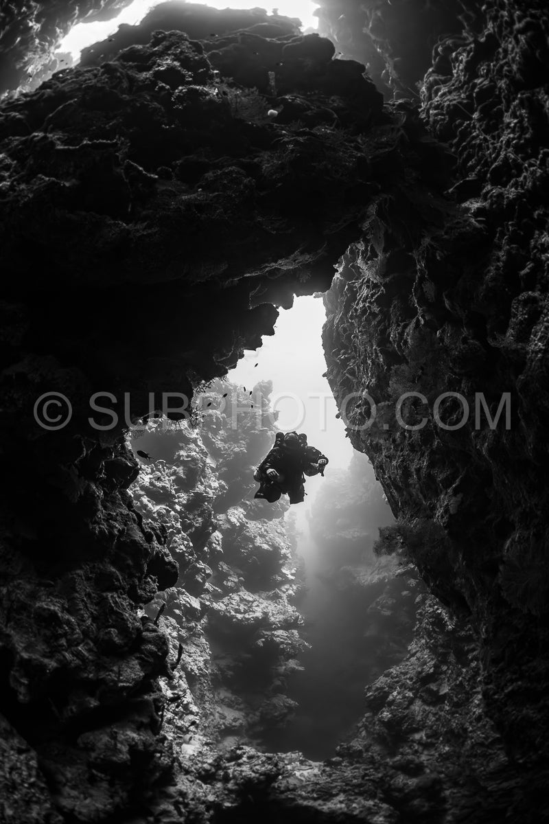 A diver wearing a rebreather swims through a narrow passage in the Red Sea. The diver is illuminated by a beam of sunlight entering the cave from above.