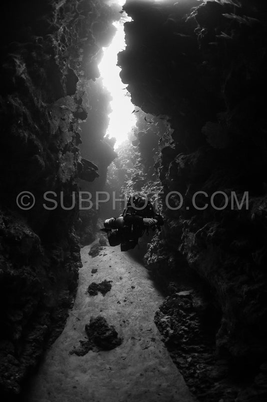 A diver- wearing a rebreather- swims through a narrow- dark passage. Light streams in from above- illuminating the diver and the rocky walls.