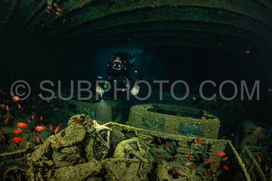 A diver wearing a rebreather swims through a dark and sunken ship in the Red Sea. The ship is covered in marine life- with small fish swimming around the diver.