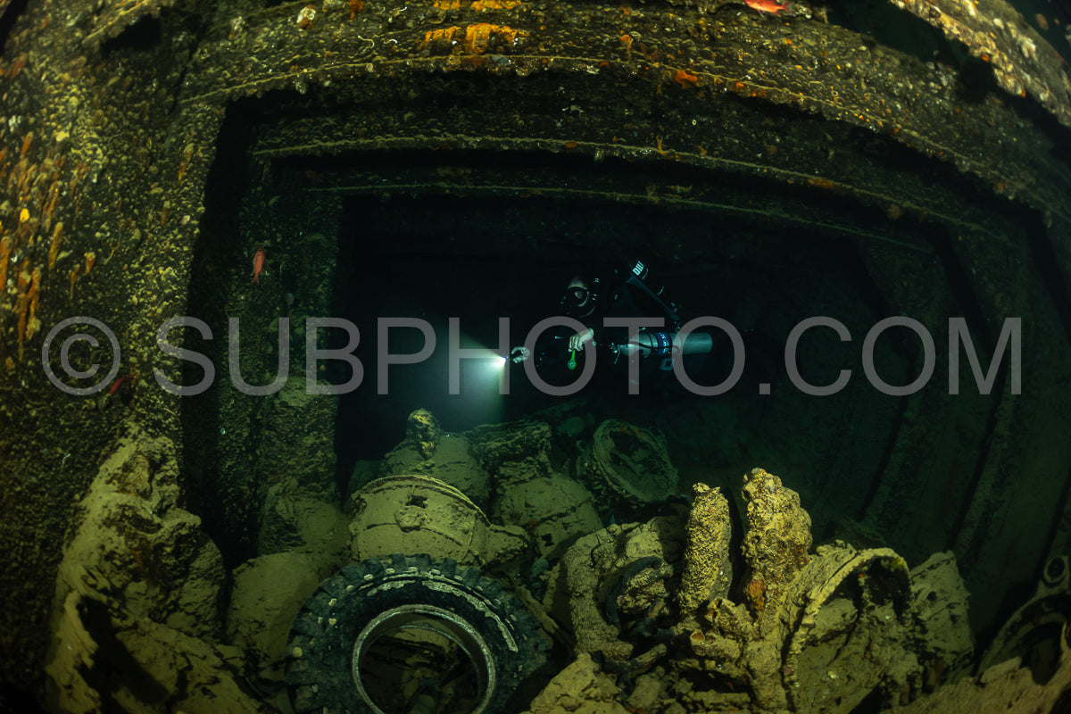 A diver is illuminated by a flashlight in the dark interior of a shipwreck- surrounded by debris and marine life. The diver appears to be using a rebreather to explore the wreck in the Red Sea.