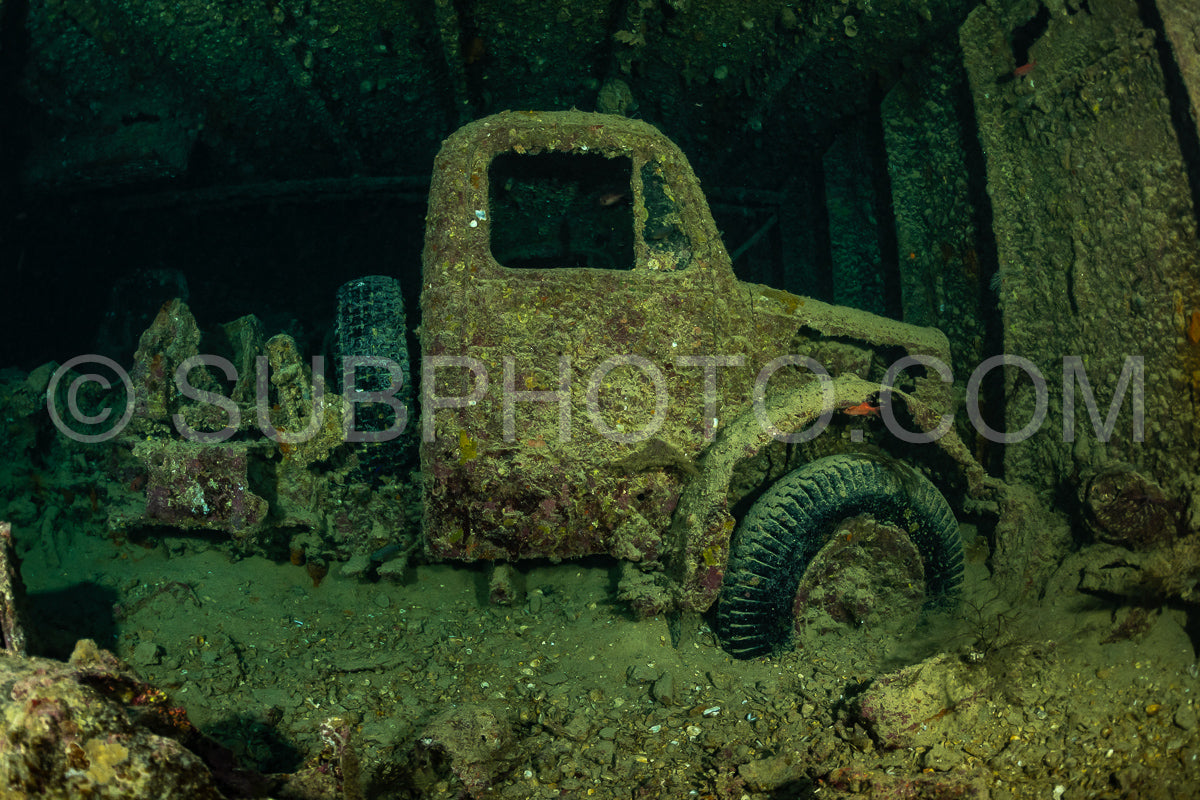 An old truck sits upright on the ocean floor- its metal frame covered in marine growth. The surrounding seafloor is a mix of sand and rubble.