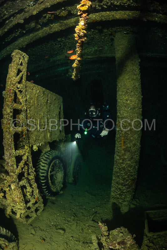 A diver- equipped with a rebreather- illuminates the interior of a shipwreck in the Red Sea. The diver's light casts a beam onto the rusting metal and marine growth covering the wreck.