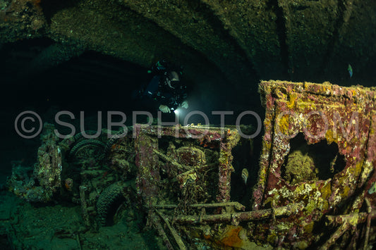 A diver using a rebreather swims through a dark underwater tunnel in the Red Sea- illuminating a rusted shipwreck with their light.