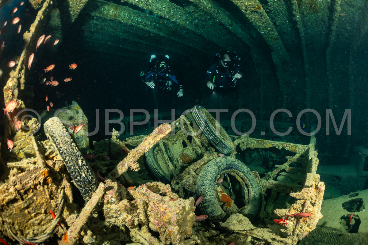 Two divers- using rebreathers- explore the interior of a shipwreck in the Red Sea. The wreck is covered in marine life and the divers are surrounded by large tires and metal debris.