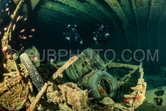 Two divers- using rebreathers- explore the interior of a shipwreck in the Red Sea. The wreck is covered in marine life and the divers are surrounded by large tires and metal debris.