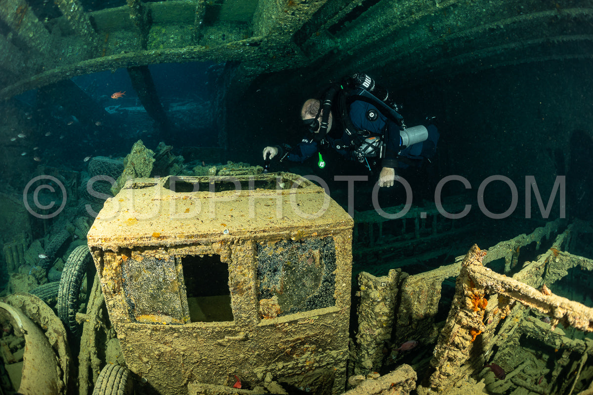 A scuba diver wearing a rebreather explores the interior of a sunken shipwreck. The diver is positioned in front of the vehicle- with the rest of the shipwreck behind them.