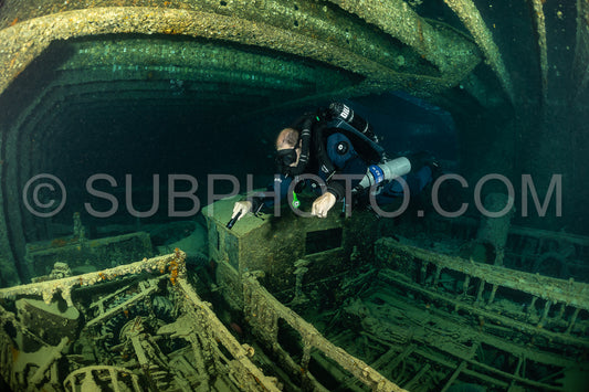 A diver wearing a rebreather swims through the interior of a shipwreck in the Red Sea. The diver is illuminated by a flashlight.