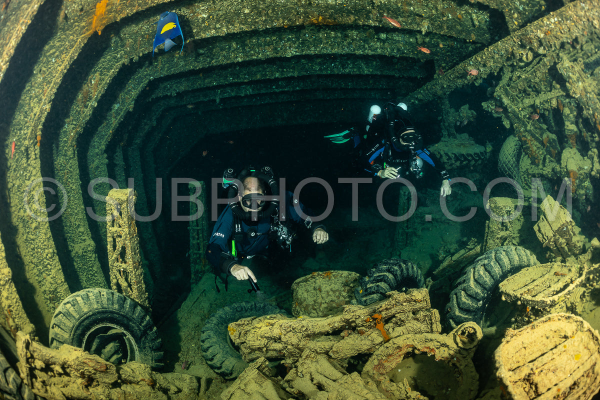 Two divers are exploring the interior of a sunken ship in the Red Sea. They are wearing rebreathers and are surrounded by the wreckage of the ship.