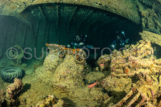 Two divers using rebreathers explore the interior of a shipwreck in the Red Sea. The ship's hull is covered in marine life and the divers are illuminated by their lights.