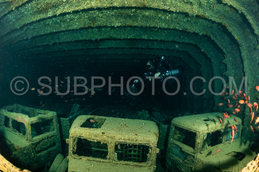 A diver wearing a rebreather swims through a large open space inside a shipwreck. The wreck is covered in algae- and the diver's light illuminates the dark interior.