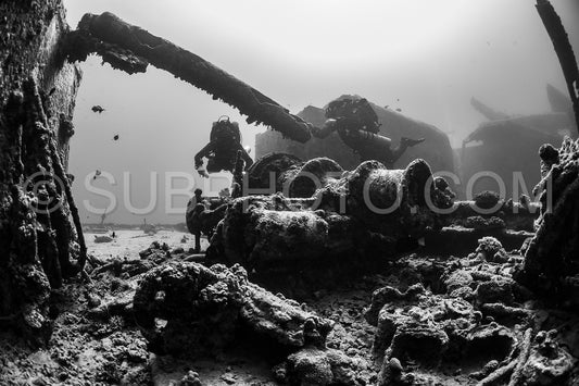 A diver with a rebreather is exploring the wreck of a ship in the Red Sea. The diver is silhouetted against the sunlight filtering through the water- and the wreck is covered in marine life.