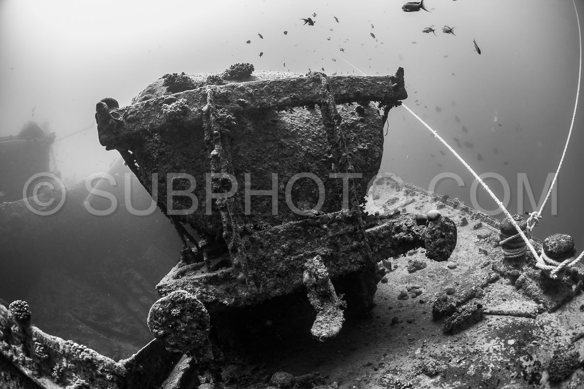 A black and white photograph of a submerged shipwreck. The metal hull is covered in barnacles and other marine life. The diver is not visible in the image but their lines can be seen.