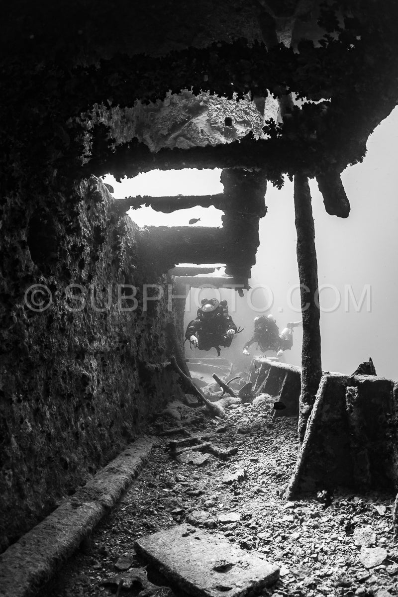 Two divers- using rebreathers- explore the interior of a shipwreck in the Red Sea. The divers are illuminated by sunlight filtering in through a broken window.